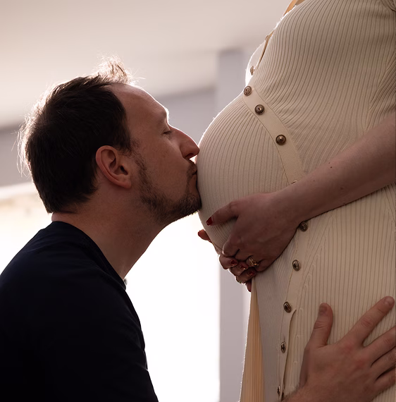 Un homme embrasse doucement le ventre d'une femme enceinte en robe crème à boutons.