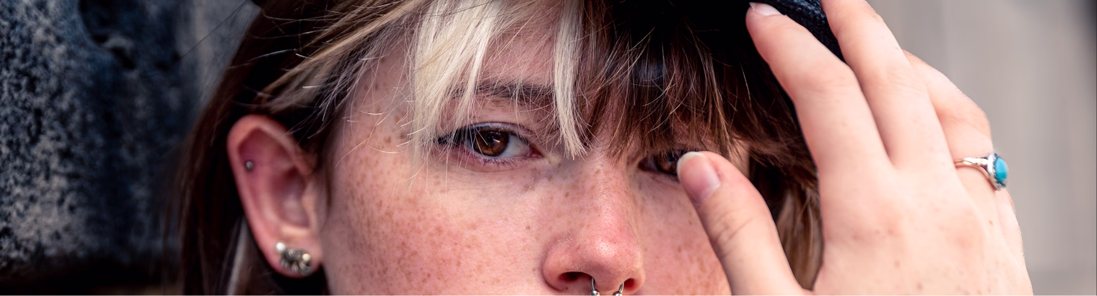 Visage rapproché d'une personne avec piercing nez, yeux marrons et cheveux bicolores touchant son chapeau.
