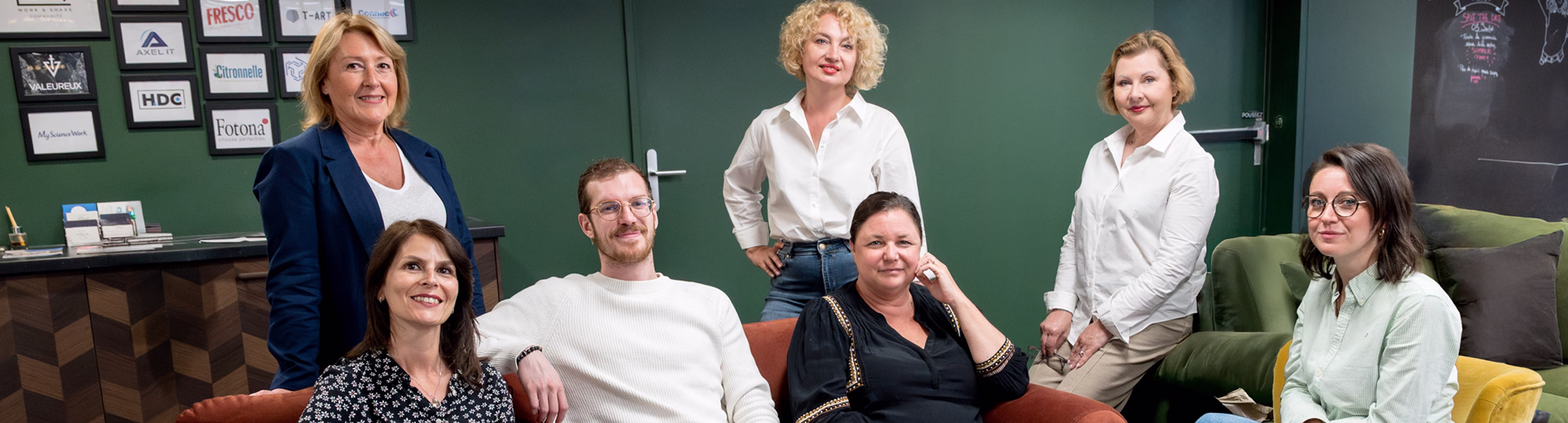 Groupe de sept personnes souriantes dans un bureau avec un mur vert et des cadres derrière eux.