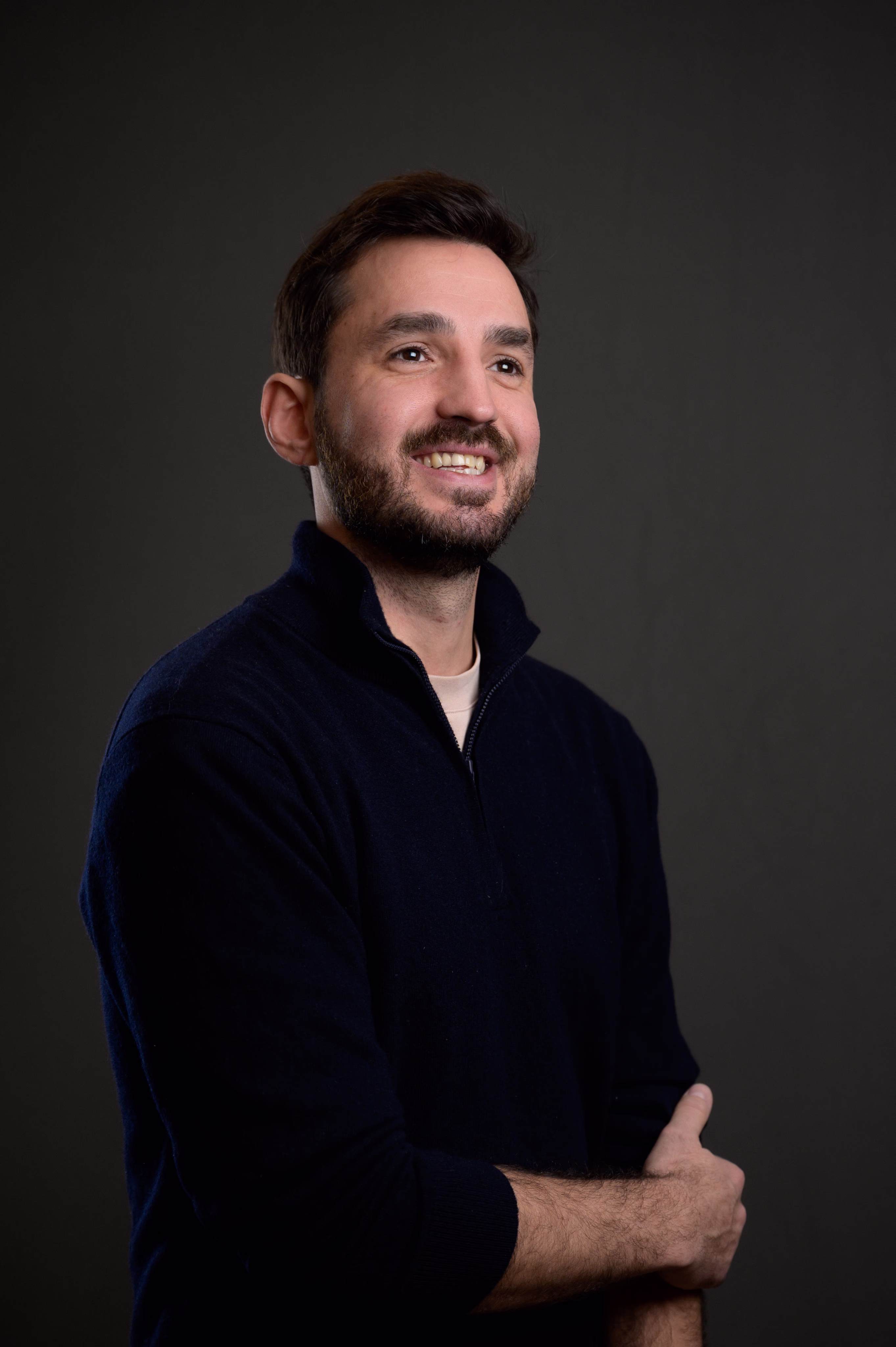 Homme souriant avec barbe, bras croisés, portant un pull bleu marine devant un fond sombre.