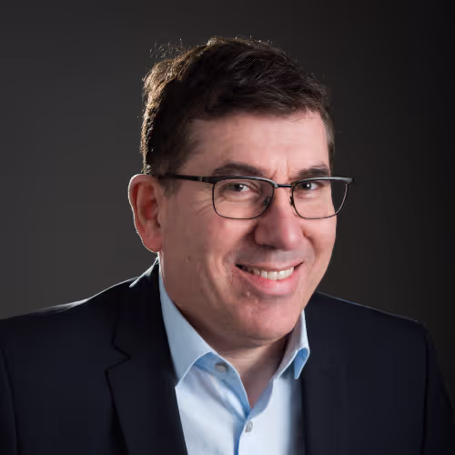 Smiling man wearing glasses, a light blue shirt, and a dark blazer against a dark background.