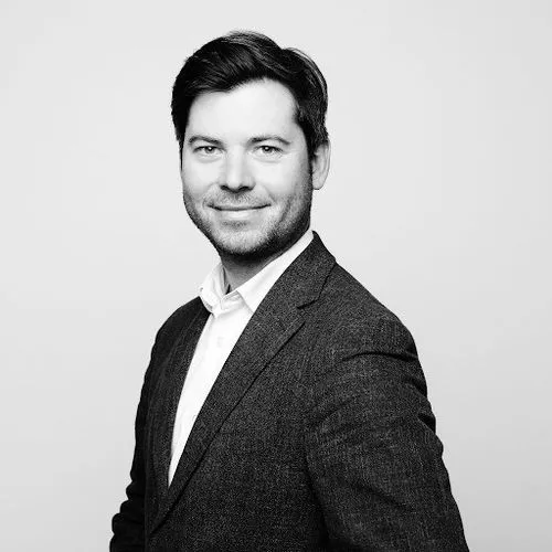 Black and white portrait of a smiling man with dark hair wearing a blazer and white shirt against a plain background.