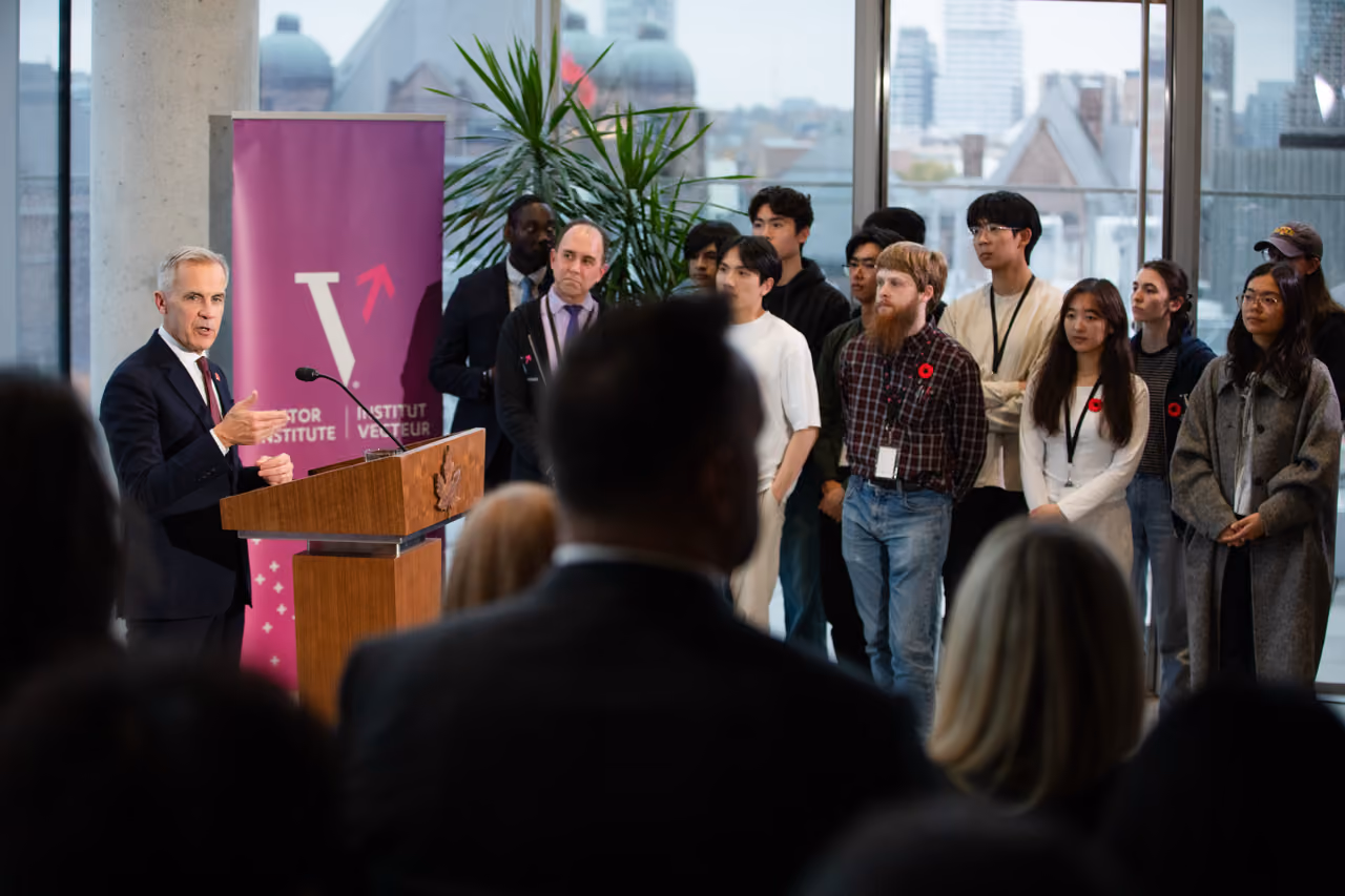 Prime Minister of Canada Mark Carney addresses an audience from a wooden podium at a Vector Institute event, with the pink Vector Institute banner visible behind him. A group of approximately a dozen young researchers and students stand attentively to his right.