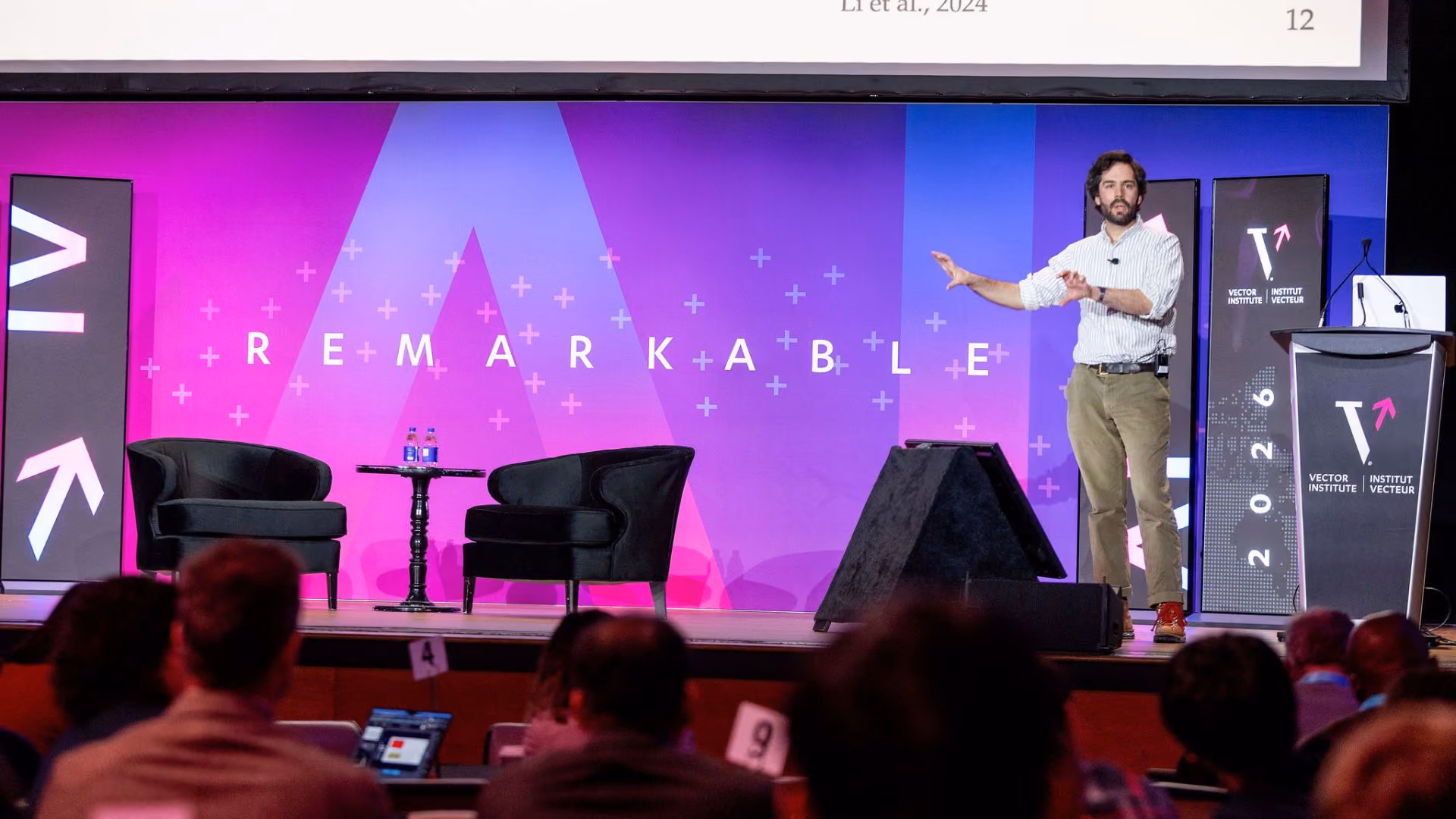 Chris J. Maddison presents on stage at Vector Institute's Remarkable event, gesturing toward the audience. A research slide is projected above on the screen behind him, with the pink and purple "Remarkable" stage backdrop and Vector Institute branding visible.