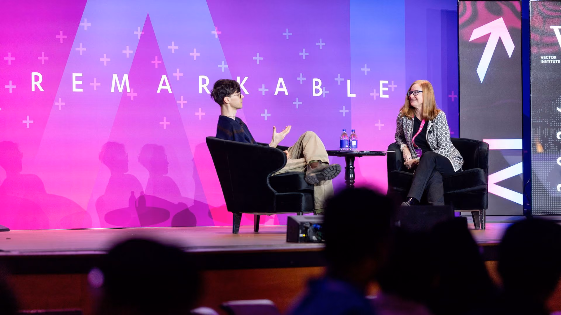 Nick Frosst and Glenda Crisp seated in armchairs on stage for a fireside chat at Vector Institute's Remarkable event, with the pink and purple "Remarkable" backdrop behind them. One gestures animatedly while the other listens and smiles.
