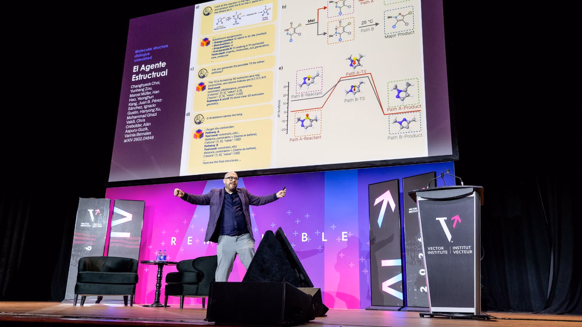 Alan Aspuru-Guzik presents on stage at Vector Institute's Remarkable event with arms spread wide, in front of a large projection screen displaying research on molecular structure dialogue titled "El Agente Estructural," showing chemical pathway diagrams. The "Remarkable" stage backdrop and Vector Institute branding are visible.