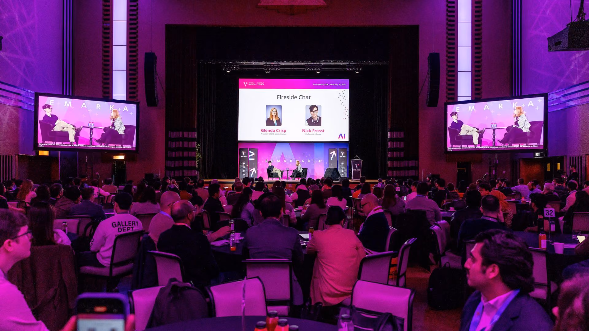 Wide shot of a packed ballroom at Vector Institute's Remarkable event. Two speakers are seated on stage for a fireside chat, with large side screens and a centre projection displaying the session title and speaker names. Hundreds of attendees are seated at round tables in purple-tinted lighting.