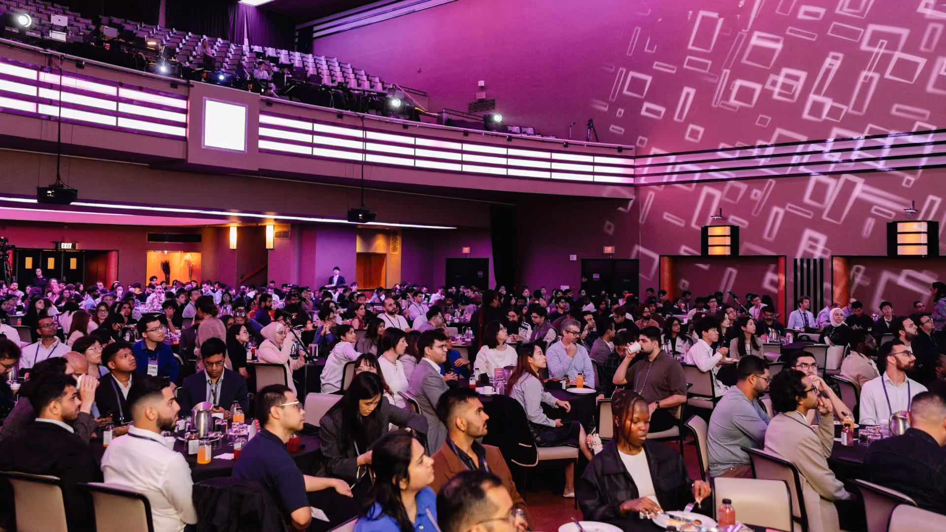 Wide shot of a packed ballroom at a Vector Institute event, with hundreds of attendees seated at round tables under dramatic purple and pink lighting. Geometric projections illuminate the walls and a mezzanine level with additional seating is visible above.