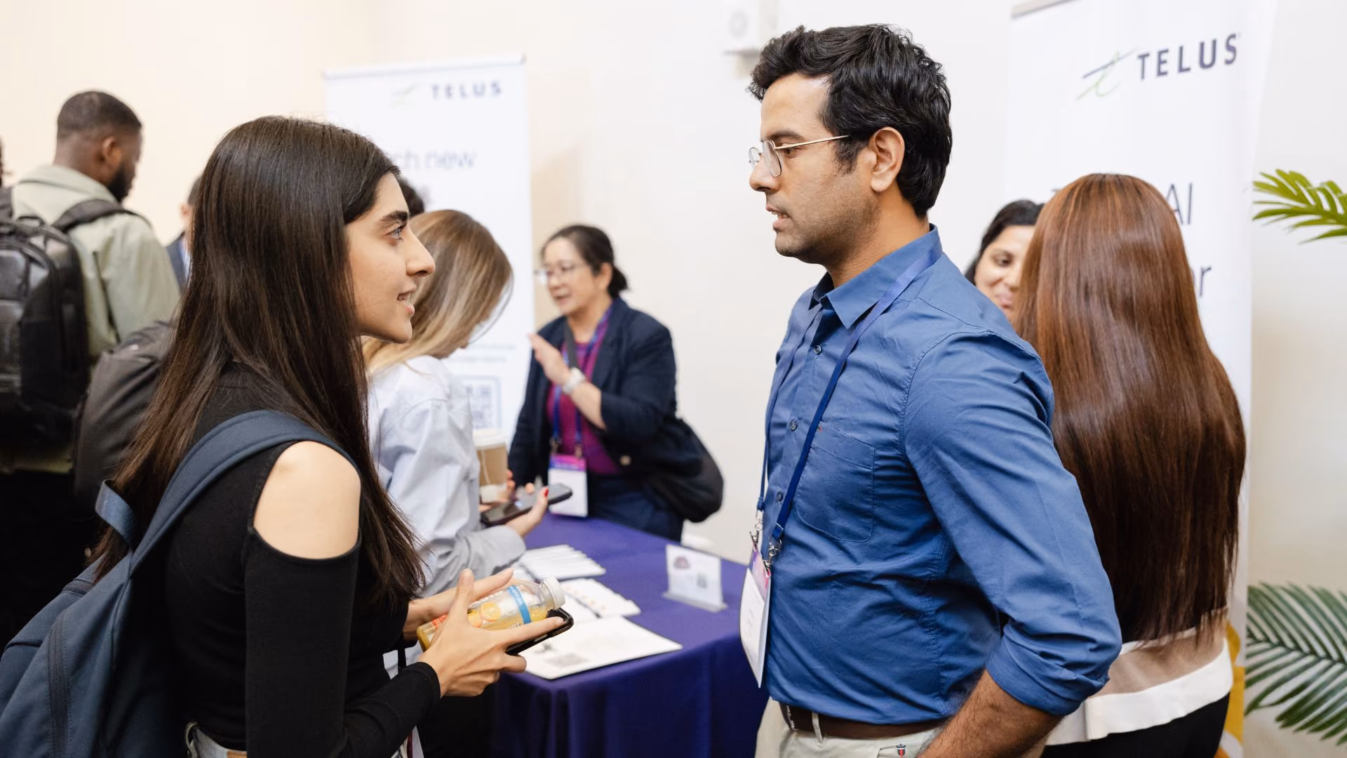 Two conference attendees engaged in conversation at a networking or career fair session. A TELUS branded booth and banner are visible in the background, with other attendees interacting nearby.