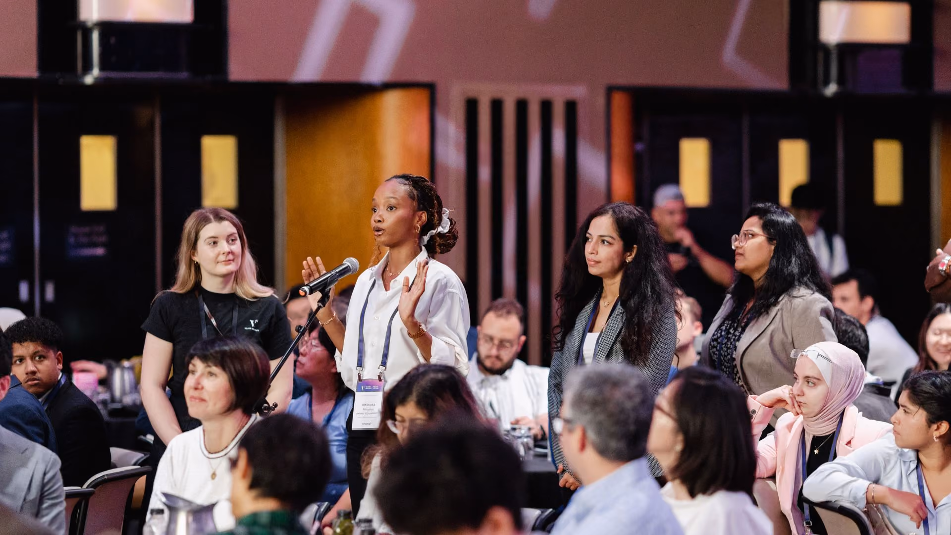 An attendee in a white blouse speaks into a microphone during a Q&A session in a large ballroom at a Vector Institute event, gesturing with both hands. A Vector Institute staff member holds the microphone stand nearby, with other standing and seated attendees visible around her.