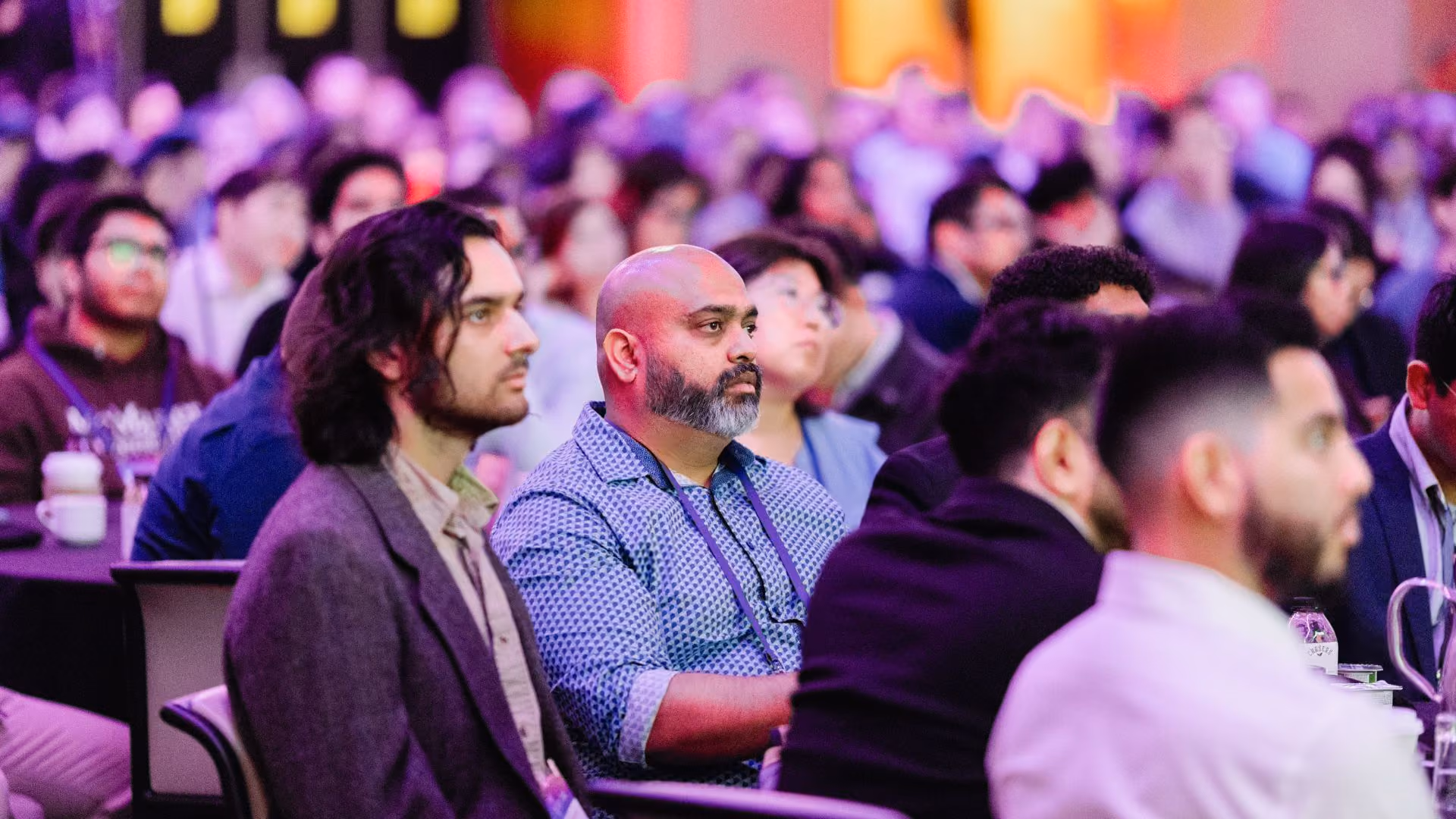 A close-up of attendees seated at round tables in a large, purple-lit ballroom, listening attentively during a session at a Vector Institute event. A man in a blue patterned shirt and lanyard is prominent in the foreground.