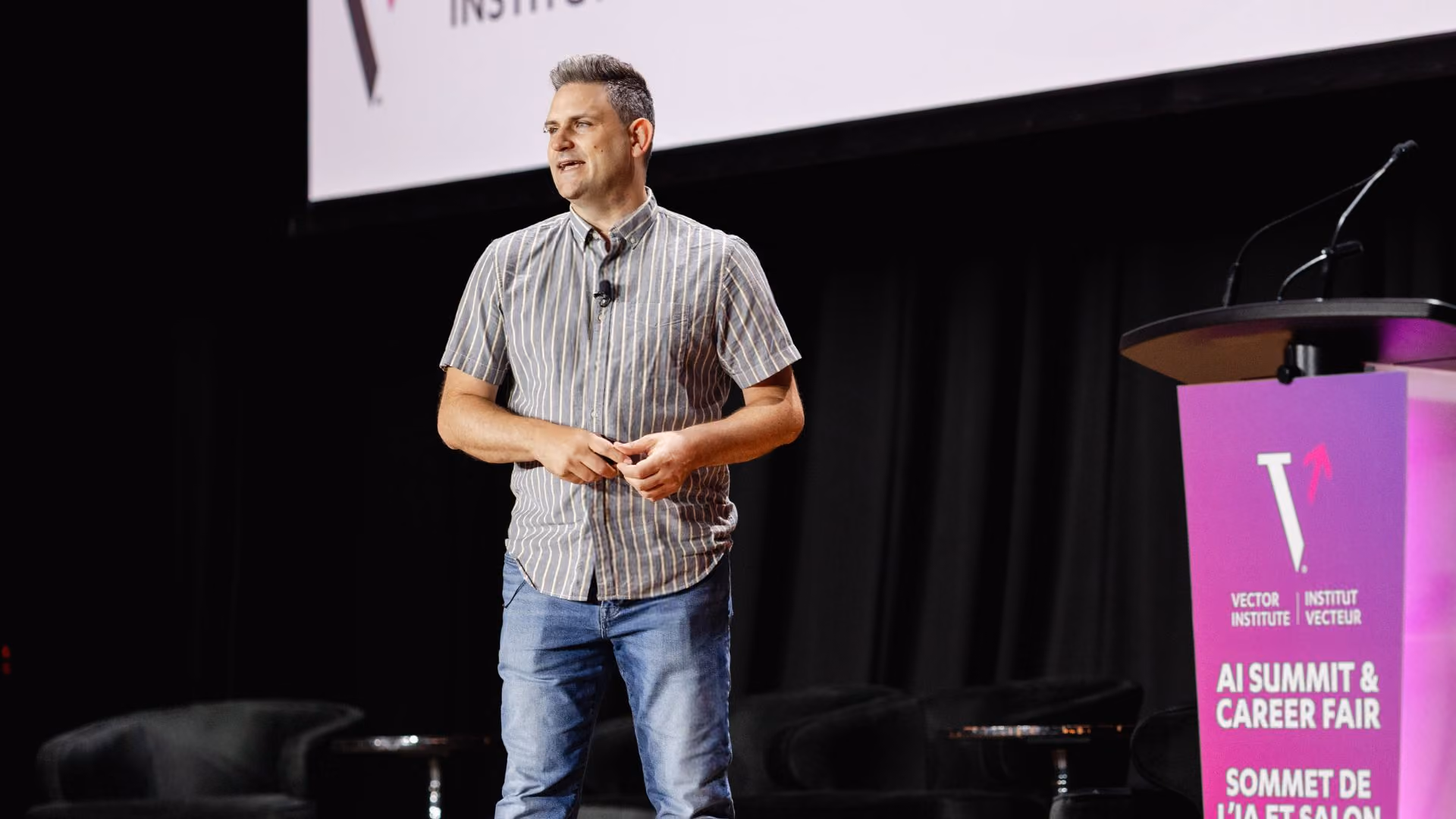 Marcus Brubaker presents on stage at the Vector Institute AI Summit and Career Fair, standing centre stage in a striped shirt with hands clasped. The Vector Institute podium is visible to his right against a dark backdrop.