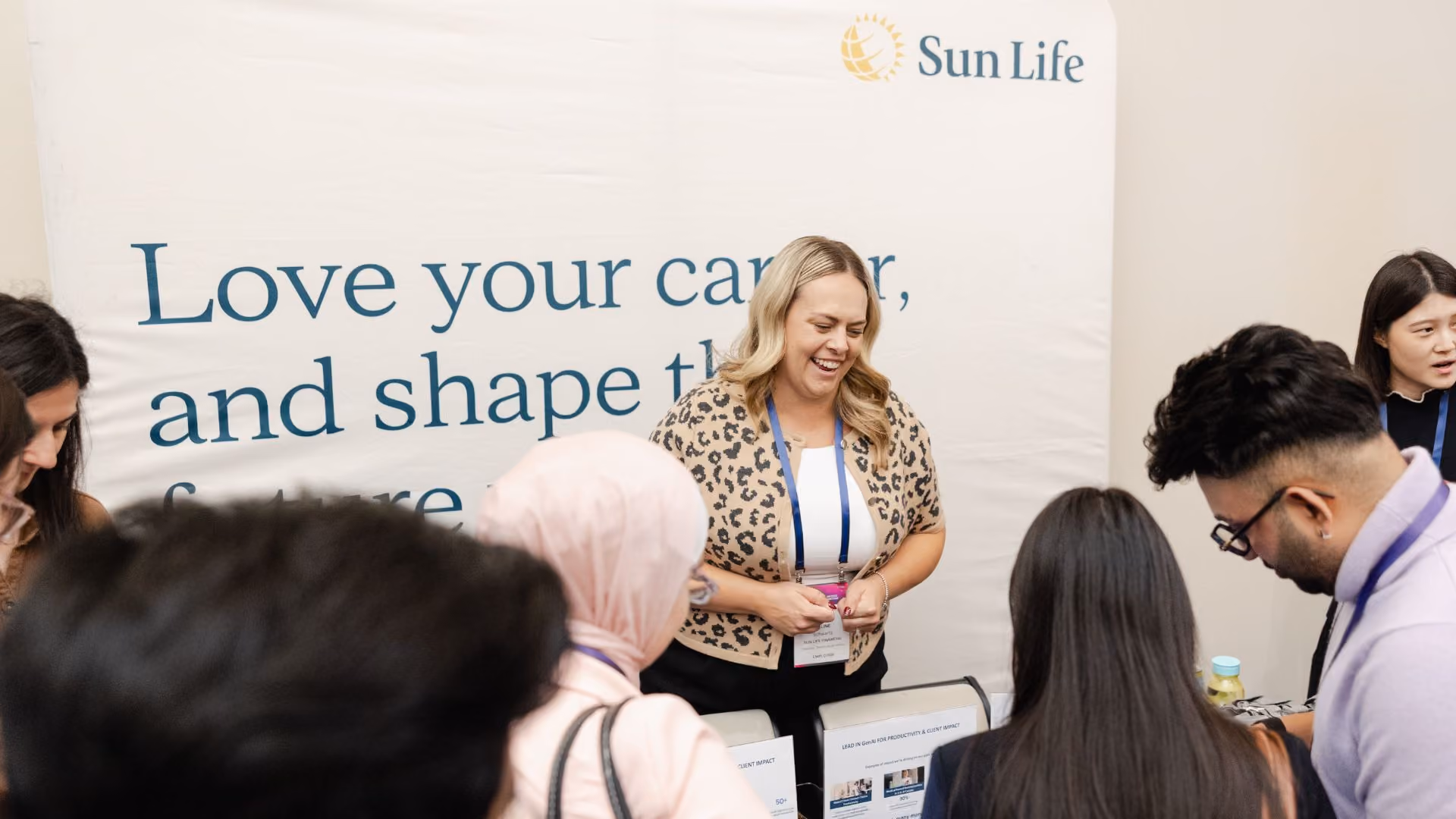 A Sun Life representative smiles and engages enthusiastically with a group of young attendees gathered around a table at a Sun Life booth. The banner behind her reads "Love your career and shape the future" with the Sun Life logo above.