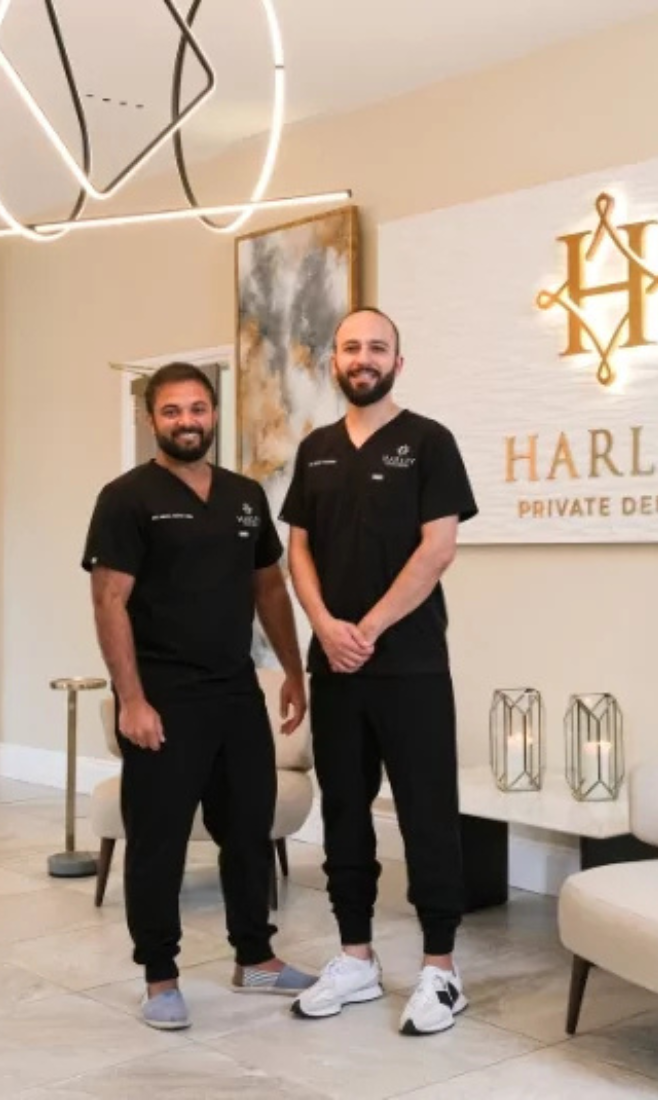 Two men in black scrubs standing and smiling inside a dental office with a sign reading 'HARL Private Dentistry' in the background.