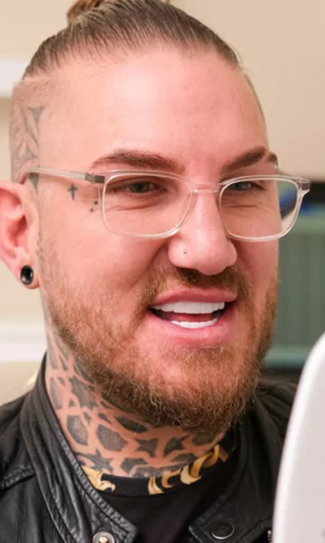 Close-up of a man with glasses, tattoos on his neck and face, a beard, and a leather jacket smiling.