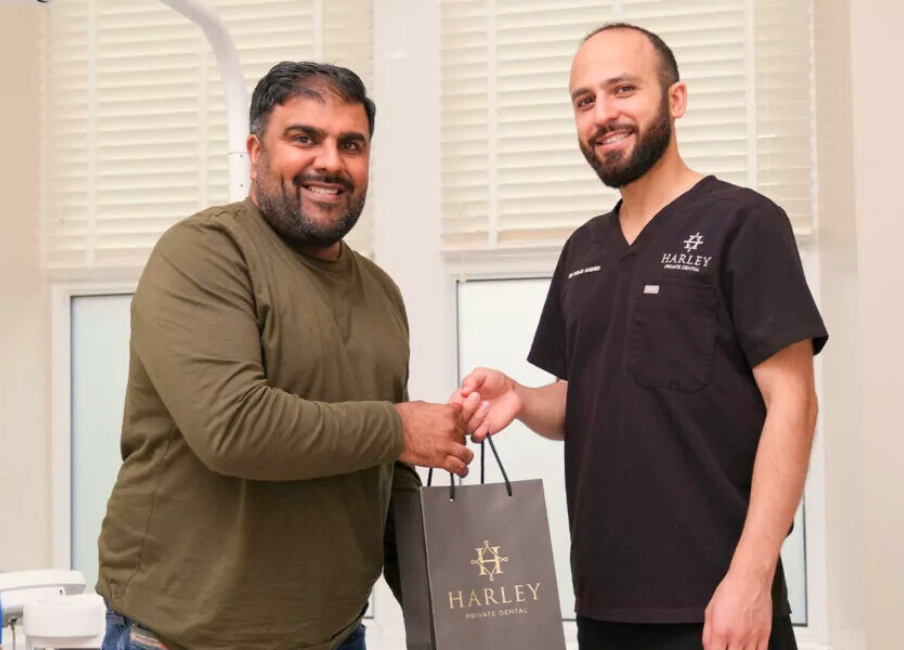A man in a green shirt receiving a gift bag from a smiling Harley Private Dental staff member in black scrubs.