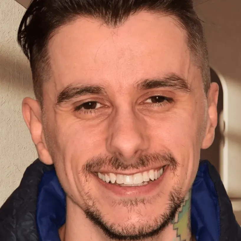 Close-up of a smiling young man with short dark hair, light facial hair, and a colorful neck tattoo.