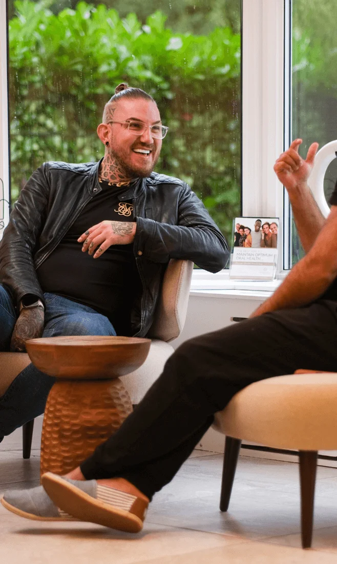 Two men sitting and conversing in a bright room with large windows and green plants outside.