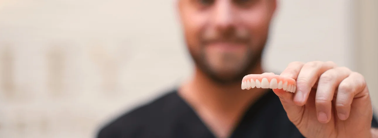 Man holding a dental prosthetic with artificial teeth, smiling in the background.