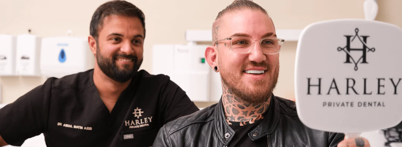 Male dental patient with tattoos smiling while looking in a handheld mirror at Harley Private Dental, with a dentist in black scrubs smiling nearby.