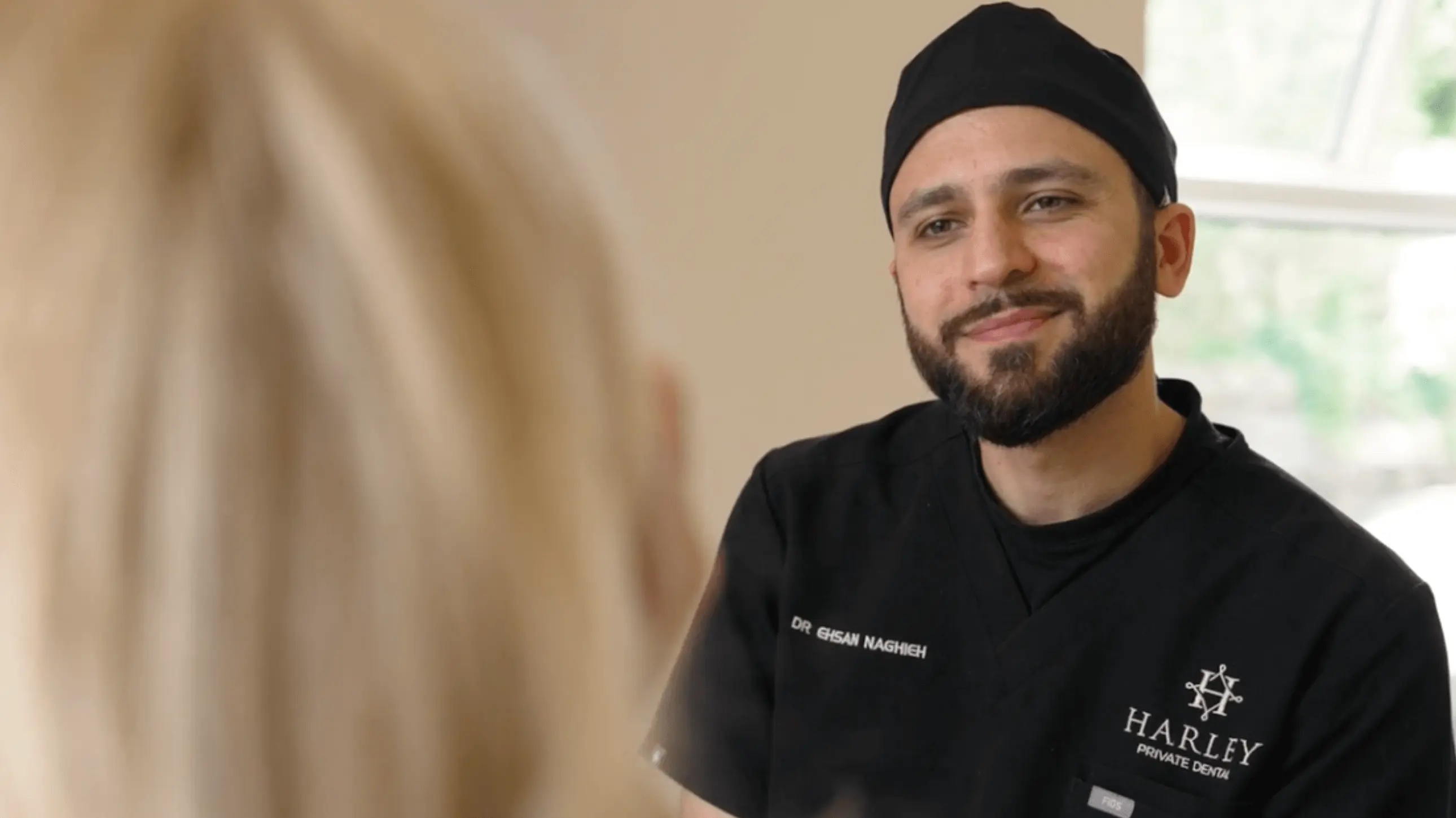 Male dentist in black scrubs with a beanie, smiling and talking to a blonde patient.