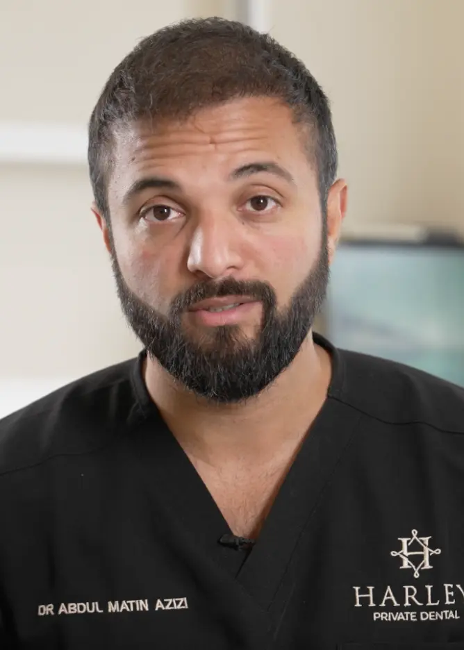 Portrait of a man with dark hair and beard wearing a black dental uniform with the name Dr Abdul Matin Azizi and Harley Private Dental logo.