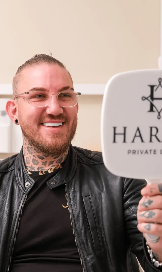 Smiling man with tattoos, piercings, glasses, and a beard wearing a black leather jacket looks at his reflection in a handheld mirror with partial text 'HAR' visible.