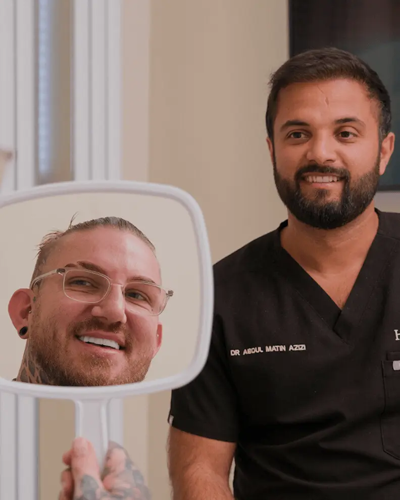 Dentist wearing black scrubs smiling next to a patient who is holding a mirror and looking at his smiling reflection showing his teeth.