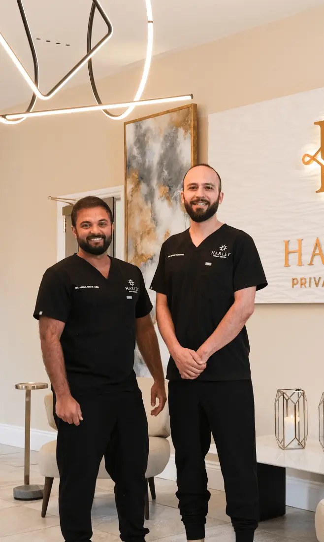 Two smiling male dentists in black scrubs standing in a modern dental clinic reception area with beige walls, abstract wall art, and a lit Harley Private Dental sign.