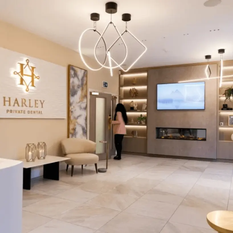 Modern dental clinic waiting area with beige walls and tile floor, featuring a lighted Harley Private Dental sign, contemporary pendant lights, beige armchair, decorative shelves with art objects, a wall-mounted TV above an electric fireplace, and a woman in a pink top opening a door.