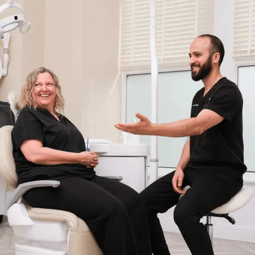 A smiling female dental patient sits in a dental chair while a male dentist in black scrubs gestures and talks to her in a bright clinic room.