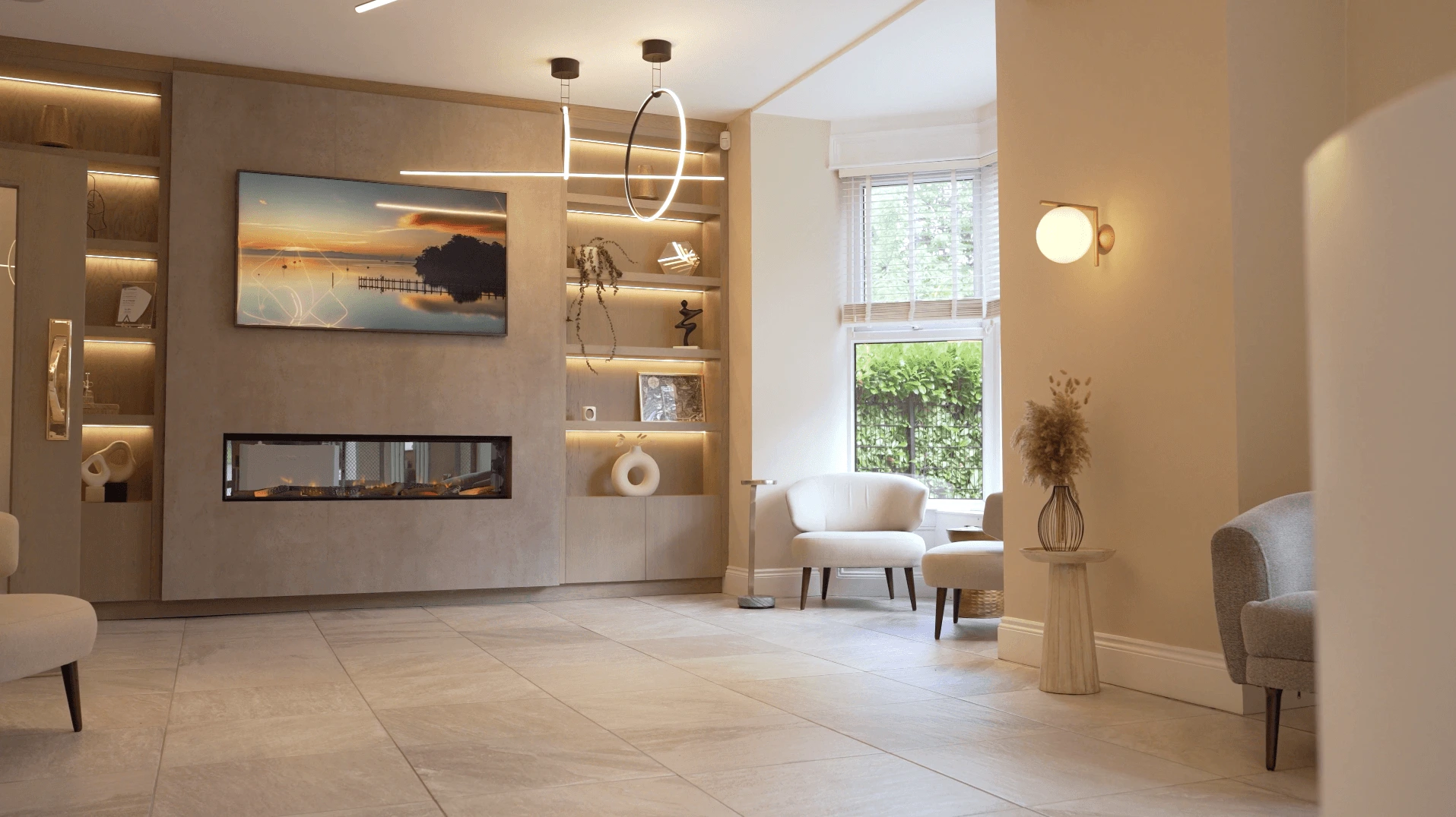 Modern, spacious living room with light beige tiled floor, built-in shelves with soft lighting, a wall-mounted TV above a rectangular fireplace, and several upholstered chairs near a window with white blinds.