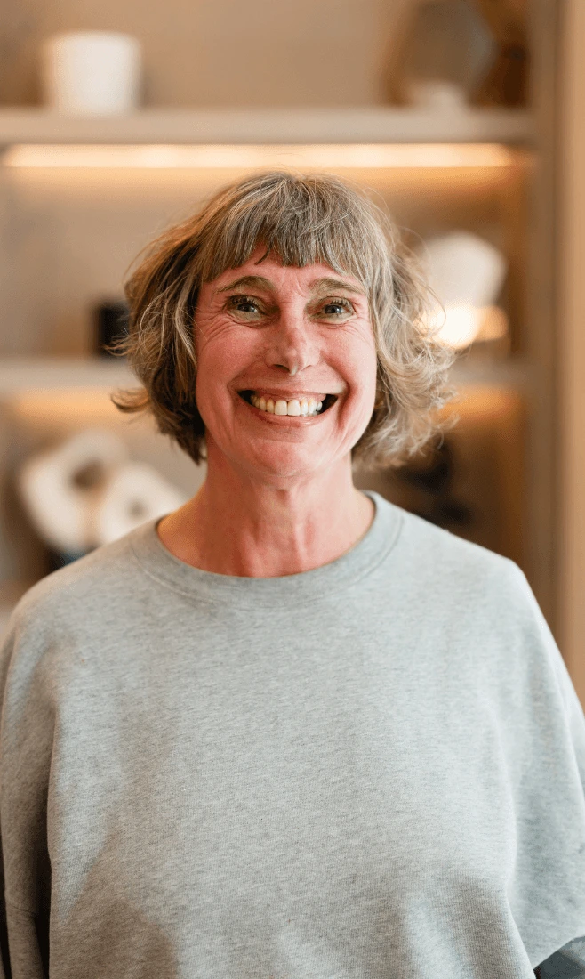 Smiling middle-aged woman with short gray hair wearing a light gray sweatshirt, standing indoors with softly lit shelves in the background.