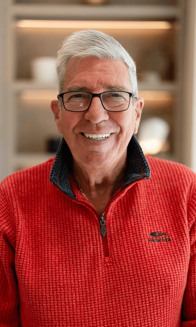 Smiling elderly man with white hair and glasses wearing a red quarter-zip sweater with a dark inner collar, standing indoors with blurred shelves in the background.
