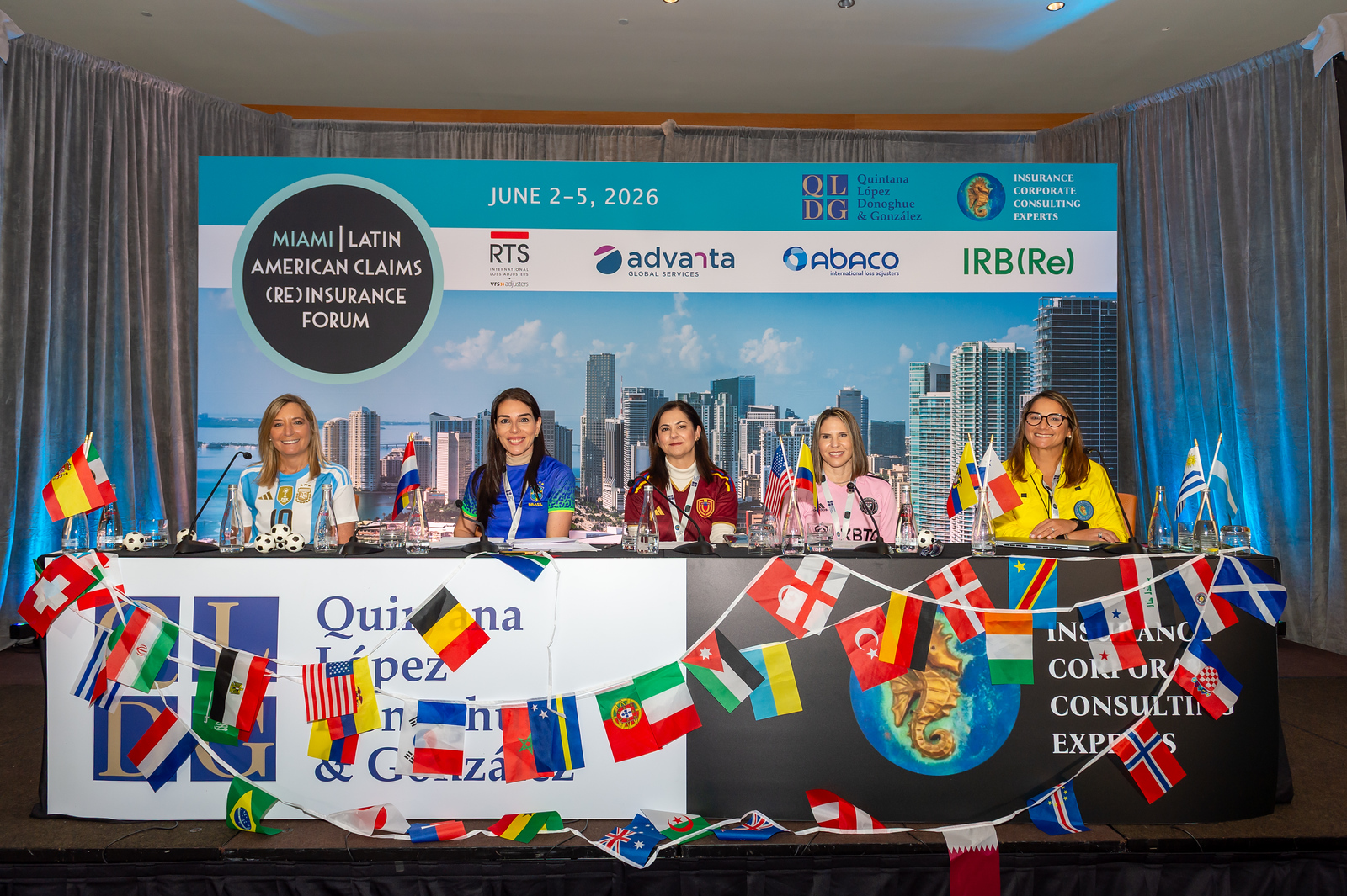 Five women sitting at a table with multiple international flags in front, participating in the Miami Latin American Claims (Re)Insurance Forum held June 2-5, 2026, with a city skyline backdrop and corporate sponsor logos above.
