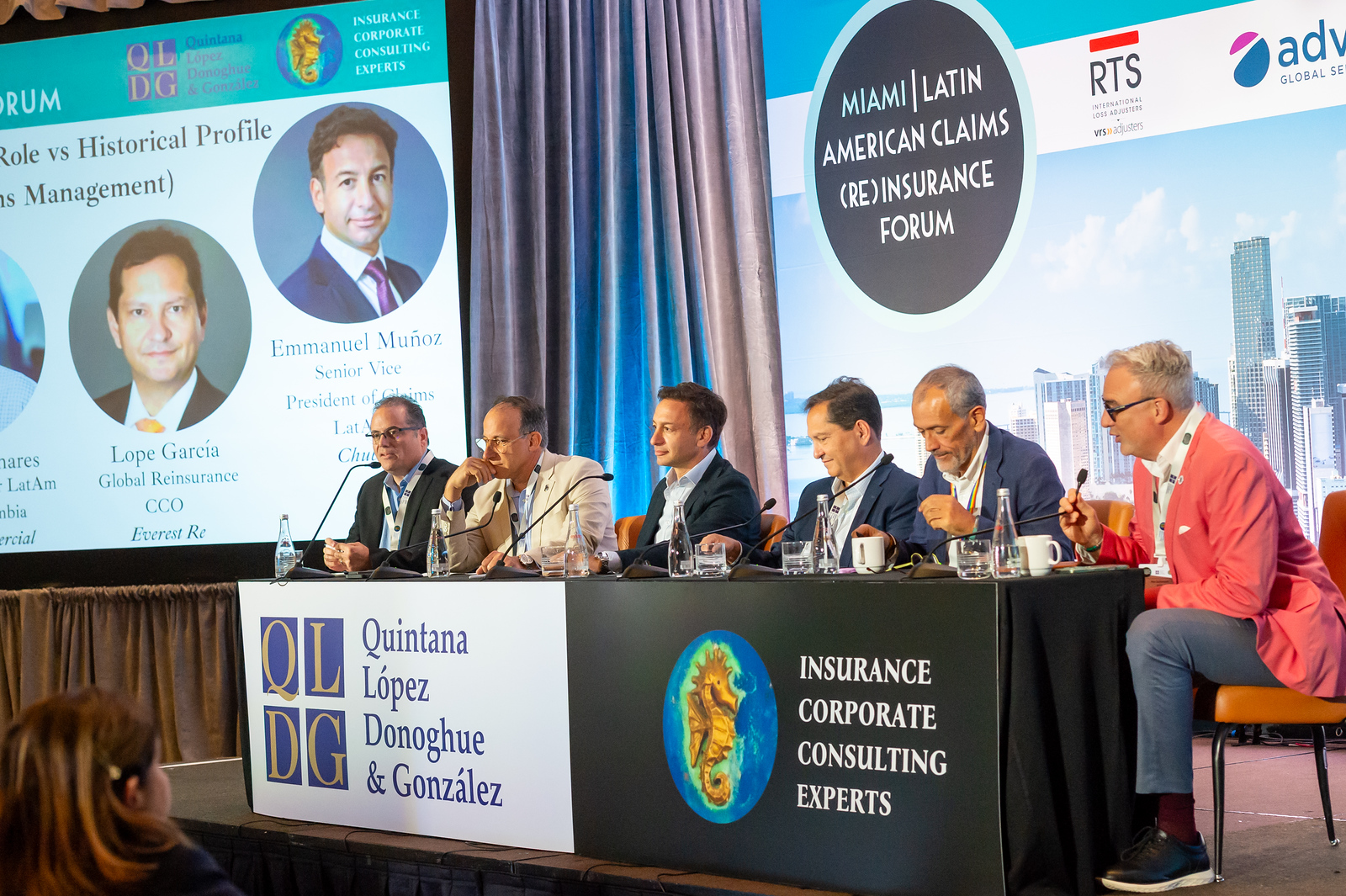 Six male panelists seated at a long table with microphones and water bottles, participating in the Miami Latin American Claims (Re)Insurance Forum, with a backdrop showing portraits of Lope García and Emmanuel Muñoz and logos for Quintana López Donoghue & González and corporate consulting experts.