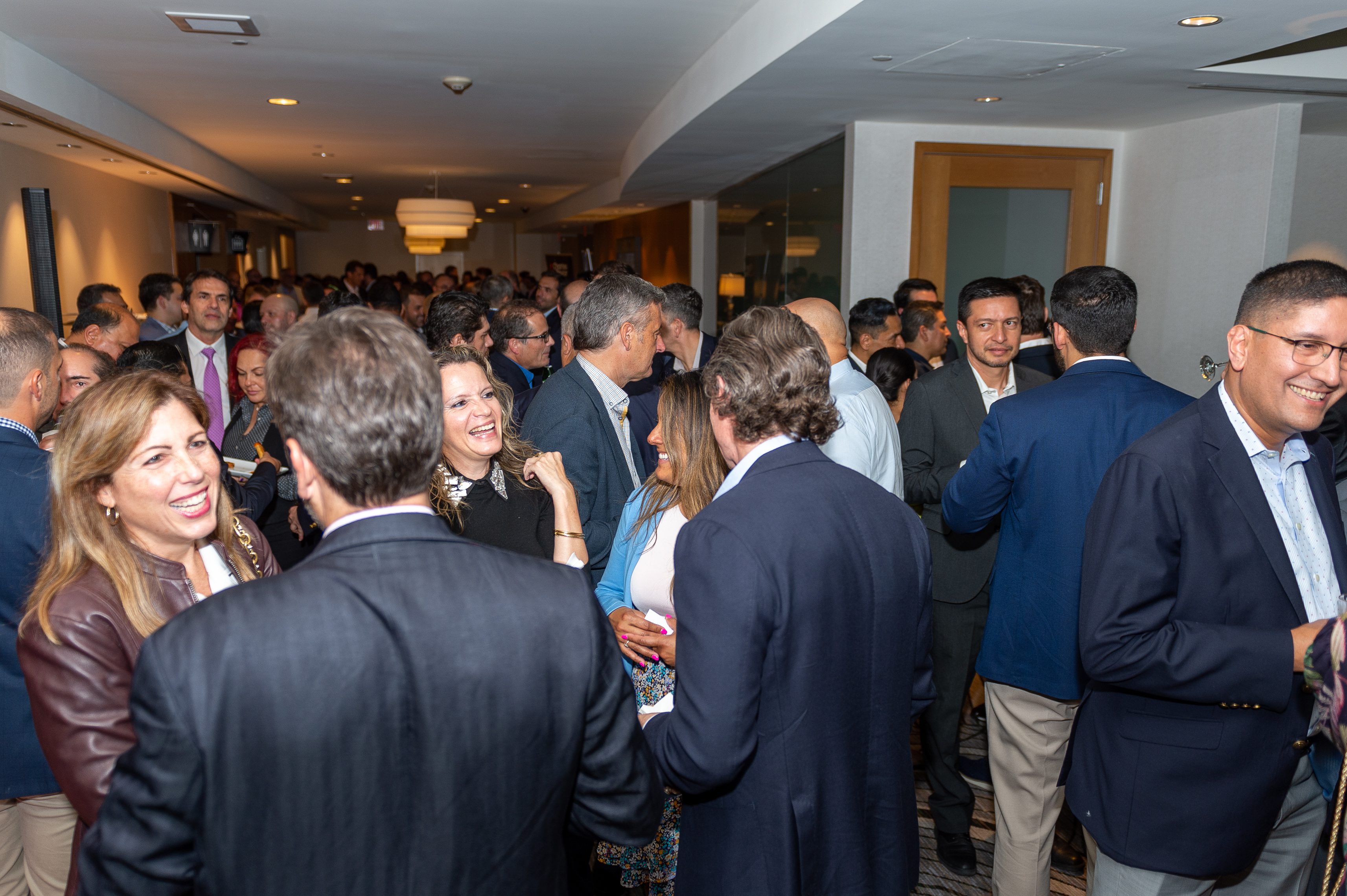 A crowded indoor social event with many people wearing business attire, engaged in conversation and smiling in a well-lit room with white walls and ceiling lights.