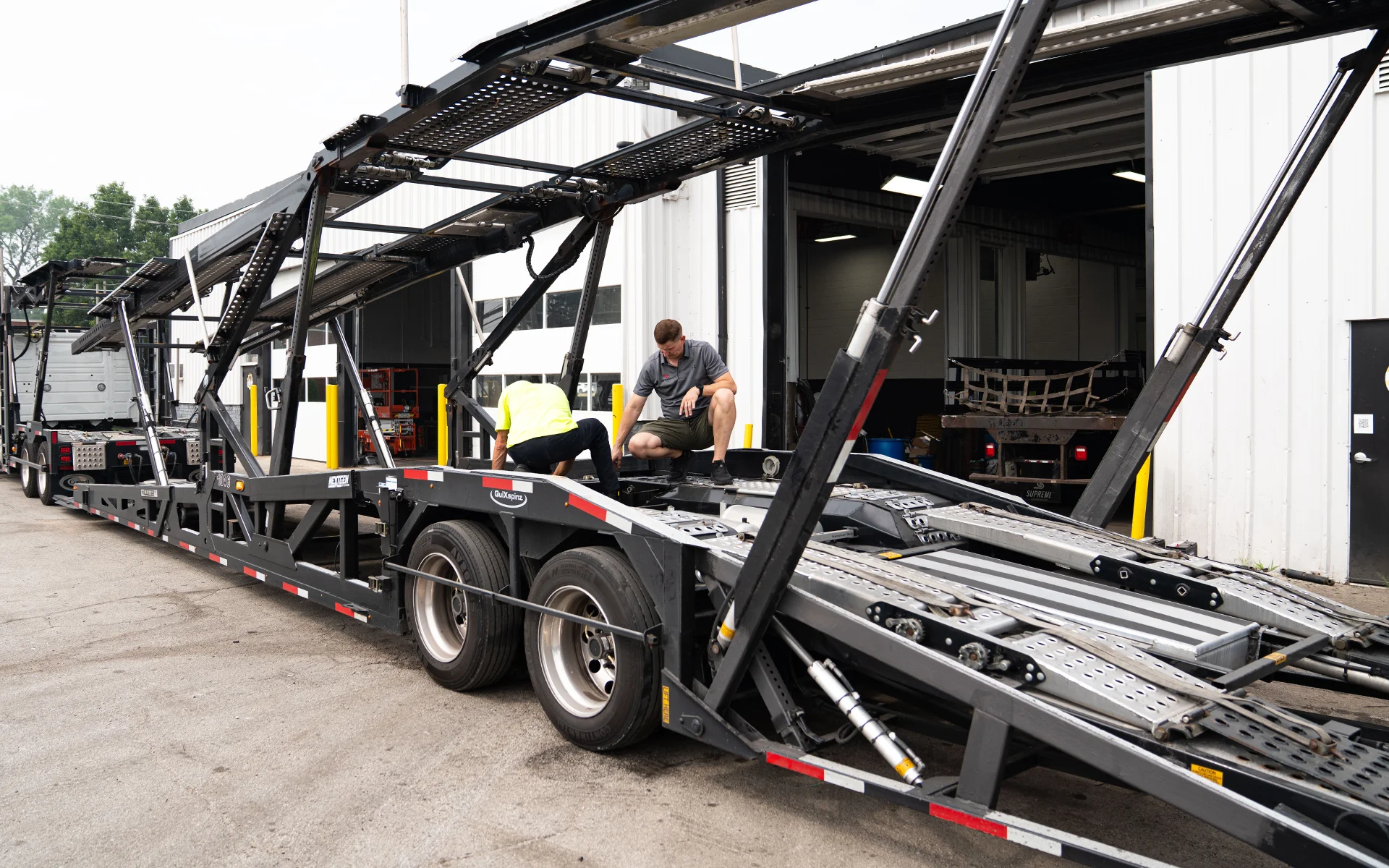 Two men working on a large car carrier trailer outside a garage with open doors.