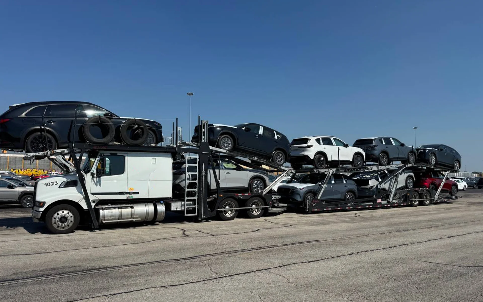 White car carrier truck loaded with multiple SUVs in a parking lot under a clear blue sky.