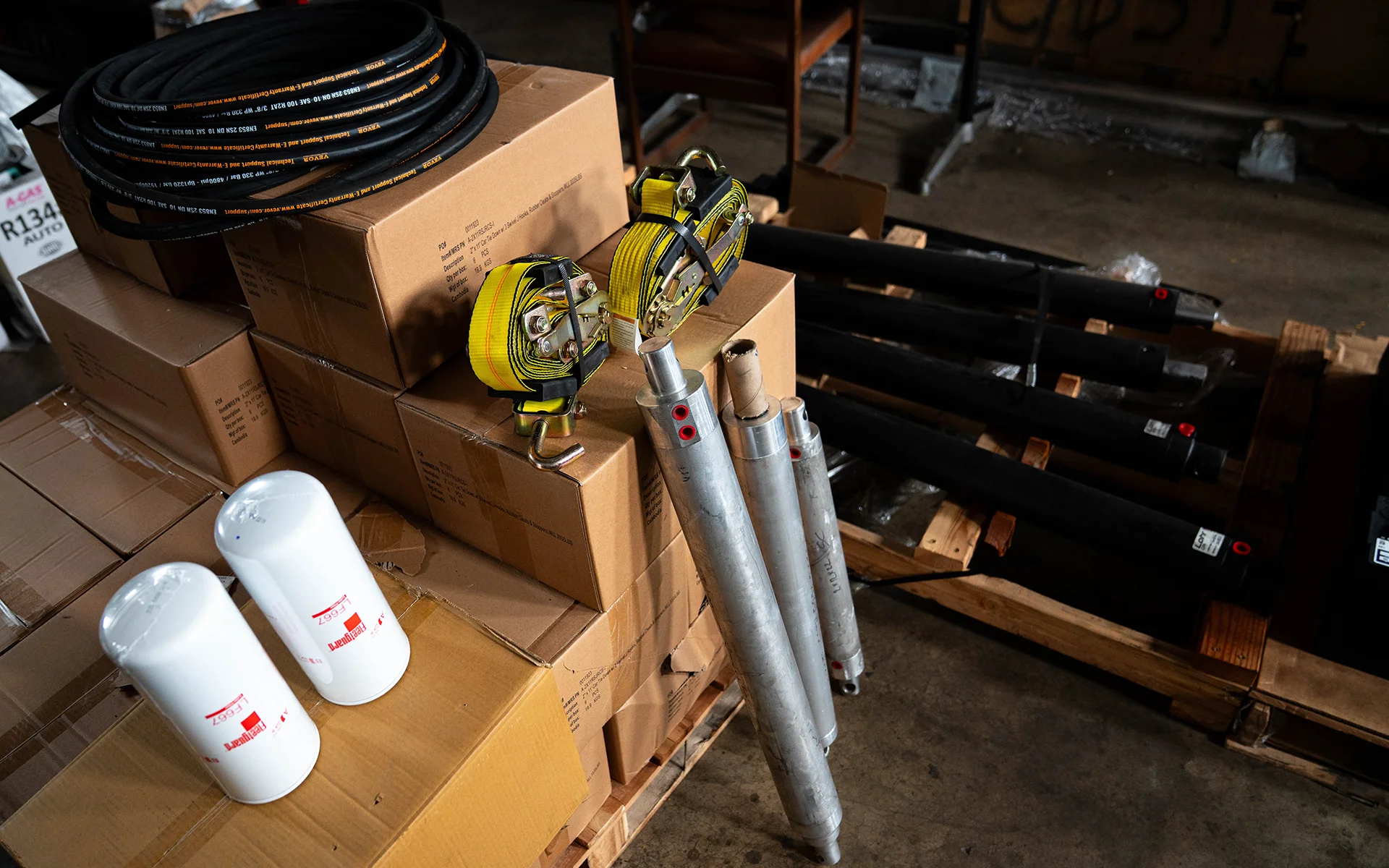 Stacked cardboard boxes with coiled hoses, yellow ratchet straps, metal cylinders, and white filters on a pallet.