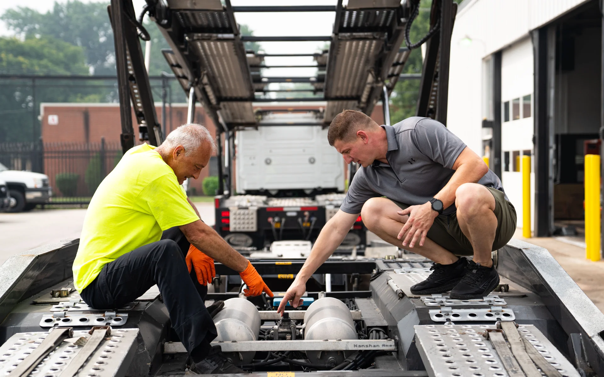 Two men inspecting mechanical parts on the back of a large car hauler outside a workshop.