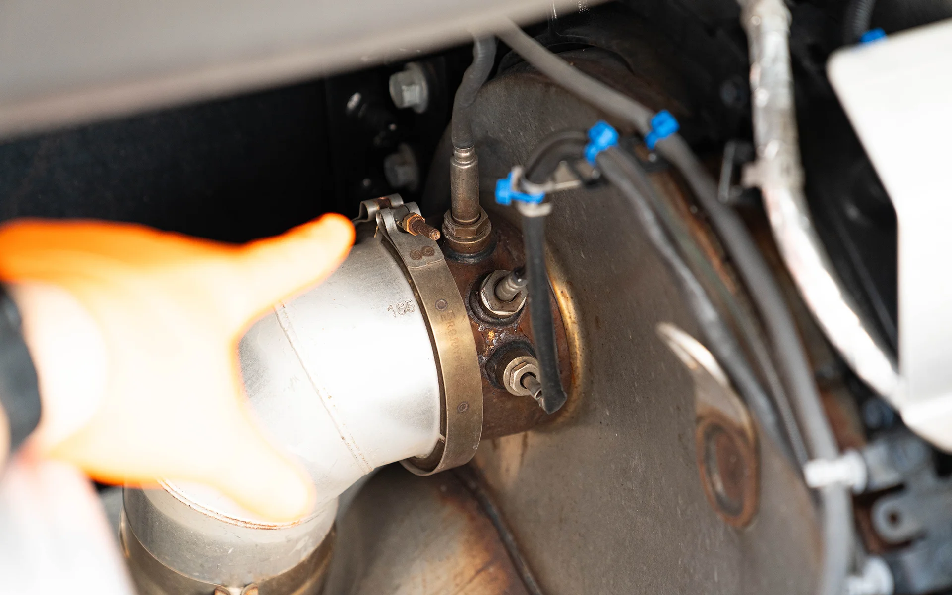 Hand wearing an orange glove pointing at a metal pipe connection with sensors and clamps in a vehicle engine.