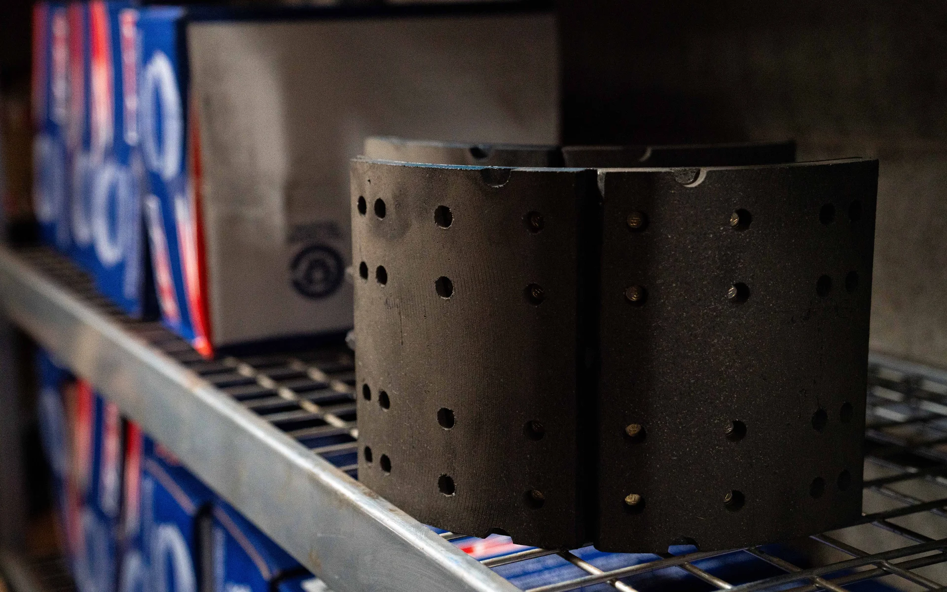 Close-up of black brake linings with holes, placed on a metal shelf with boxes in the background.