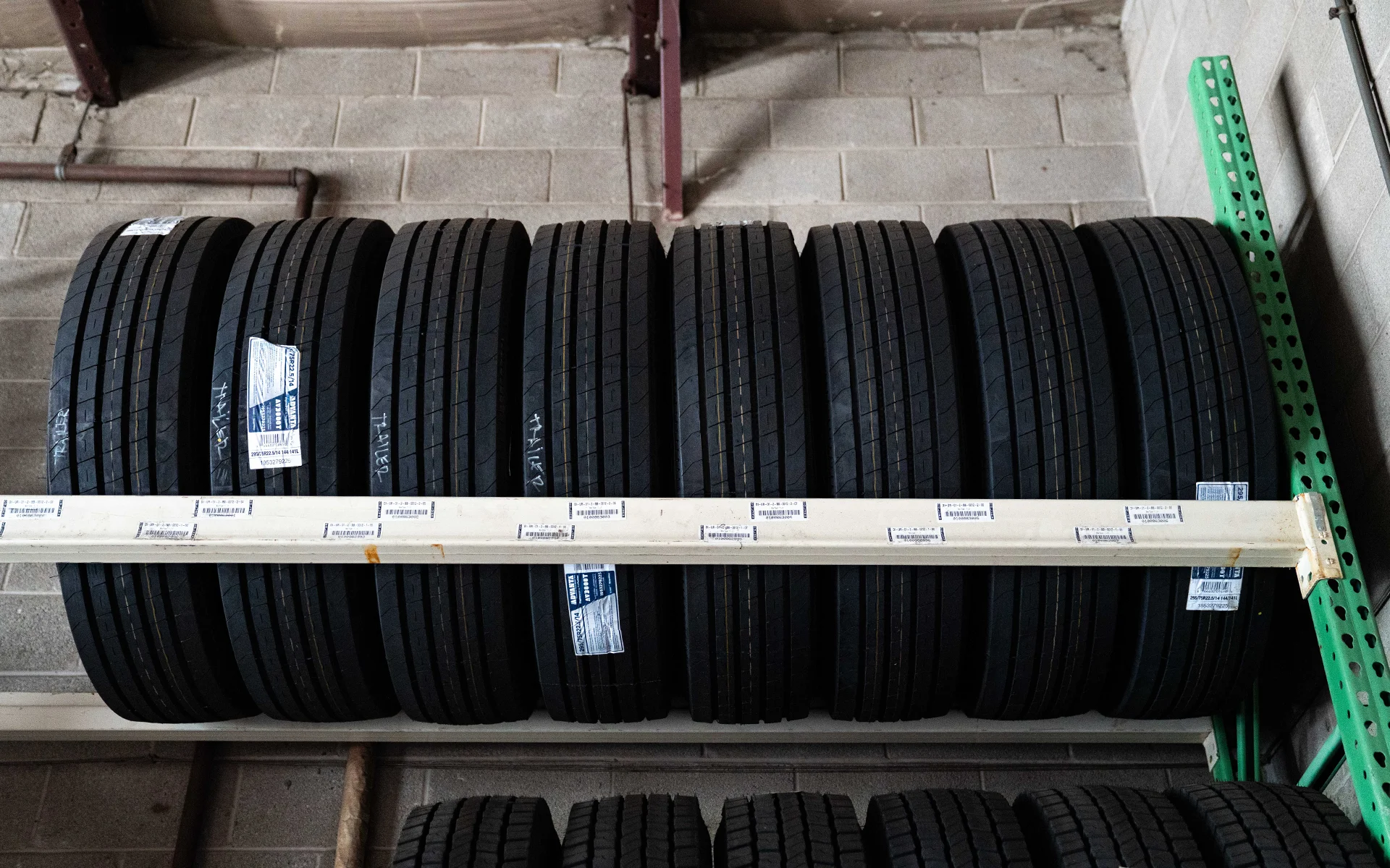 A row of nine large black tires stacked vertically on a warehouse metal rack against a concrete wall.