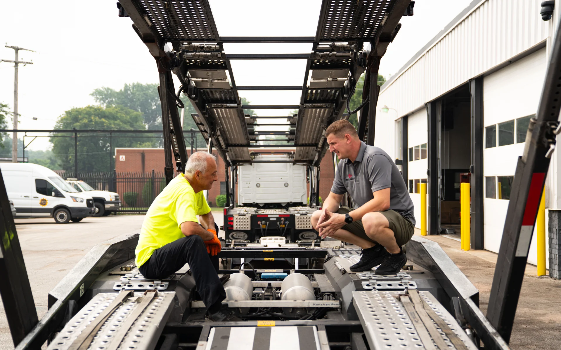 Two men talking while sitting and crouching on a car carrier trailer in an industrial area.