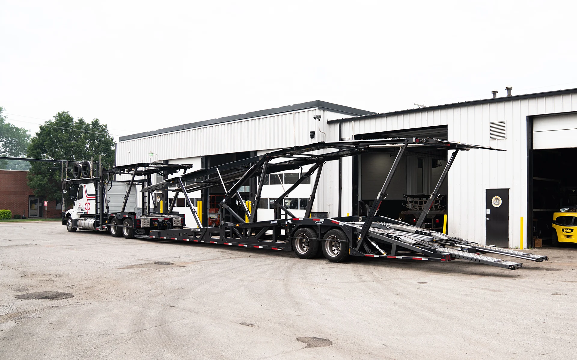 Empty black and white car carrier trailer attached to a truck outside warehouse with open garage doors.