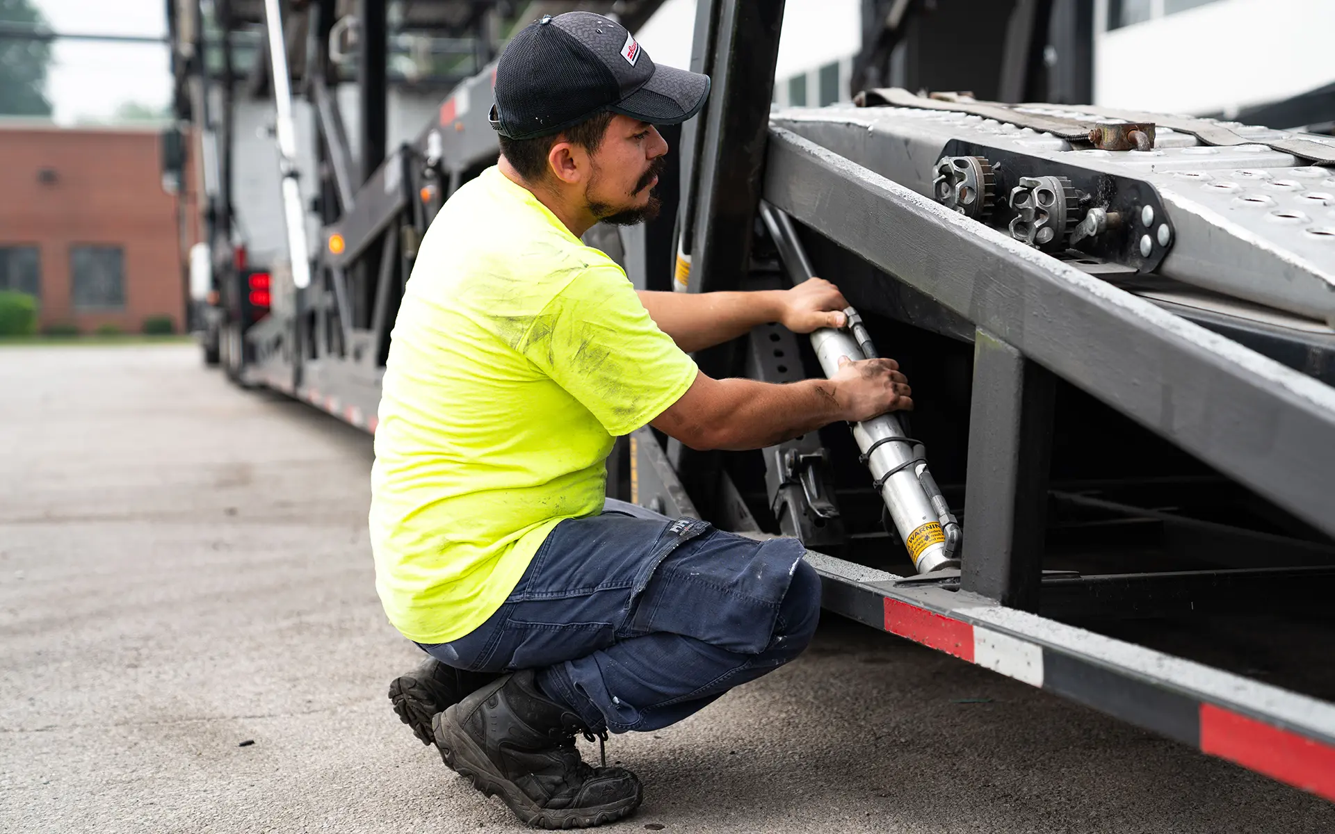 Technician squatting while inspecting or repairing a part under a large vehicle trailer.