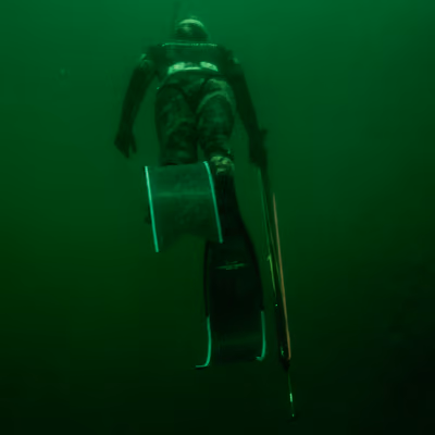 Diver in camouflage wetsuit descending in greenish water holding a spearfishing gun and fins.