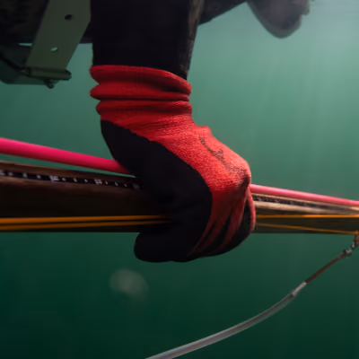Gloved hand gripping a speargun underwater with sunlight filtering through water.