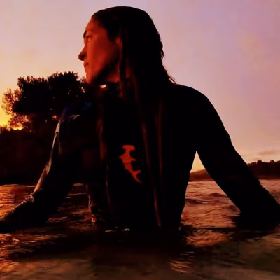 Person in a wetsuit sitting on a surfboard in water at sunset, looking left.